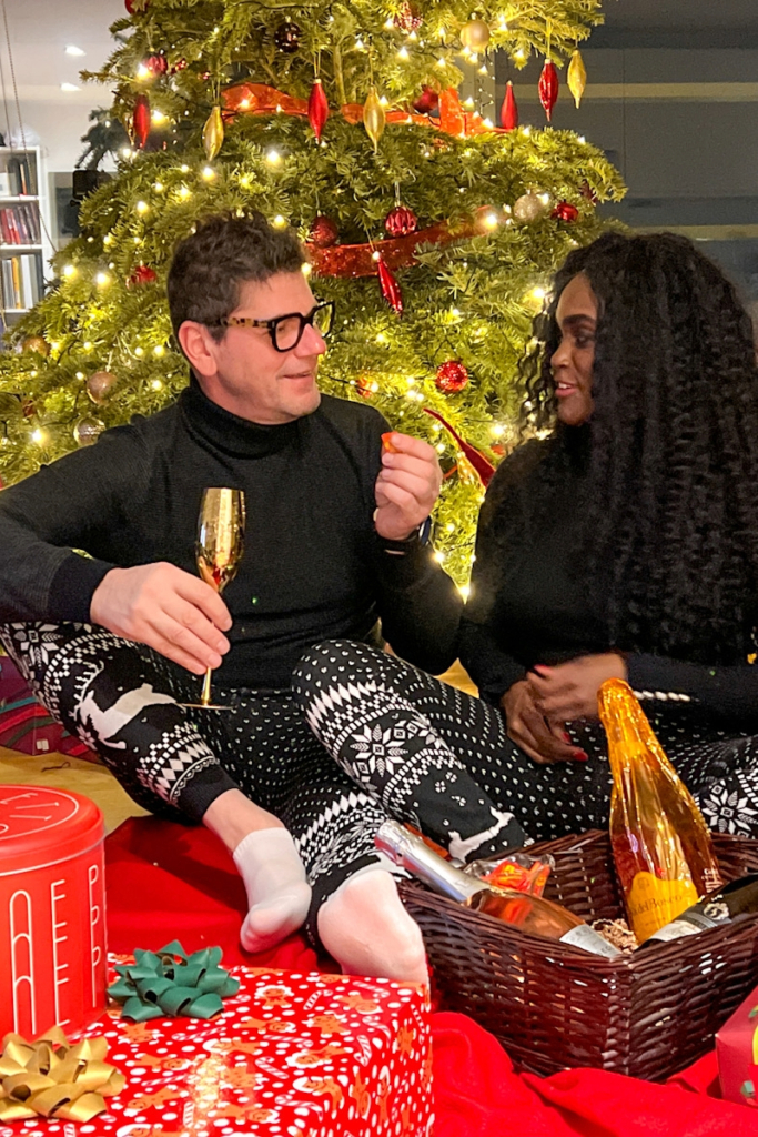 Couple sitting by a decorated Christmas tree wearing matching Christmas pyjamas at home during the festive season.