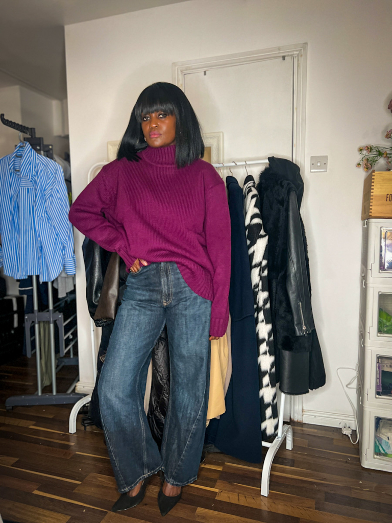 Plum turtleneck outfit featuring a fitted knit styled with dark wide-leg balloon denim and black heels beside a clothing rail.