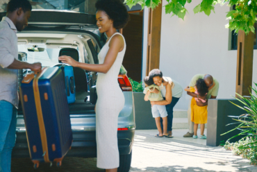 Parents load suitcases into a car while their children excitedly greet their grandparents, showing a multigenerational family travel moment before a road trip.
