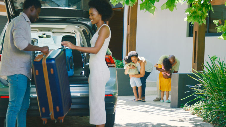 Parents load suitcases into a car while their children excitedly greet their grandparents, showing a multigenerational family travel moment before a road trip.