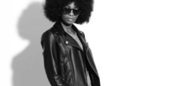 Black and white portrait of a Black woman with an afro wearing dark sunglasses, a leather jacket, a T-shirt and jeans, standing confidently and looking away from the camera, reflecting why January is such an awkward month for fashion.