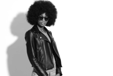 Black and white portrait of a Black woman with an afro wearing dark sunglasses, a leather jacket, a T-shirt and jeans, standing confidently and looking away from the camera, reflecting why January is such an awkward month for fashion.