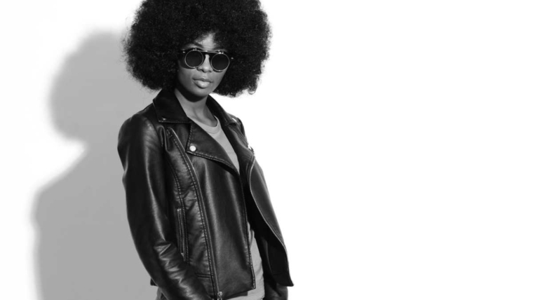 Black and white portrait of a Black woman with an afro wearing dark sunglasses, a leather jacket, a T-shirt and jeans, standing confidently and looking away from the camera, reflecting why January is such an awkward month for fashion.