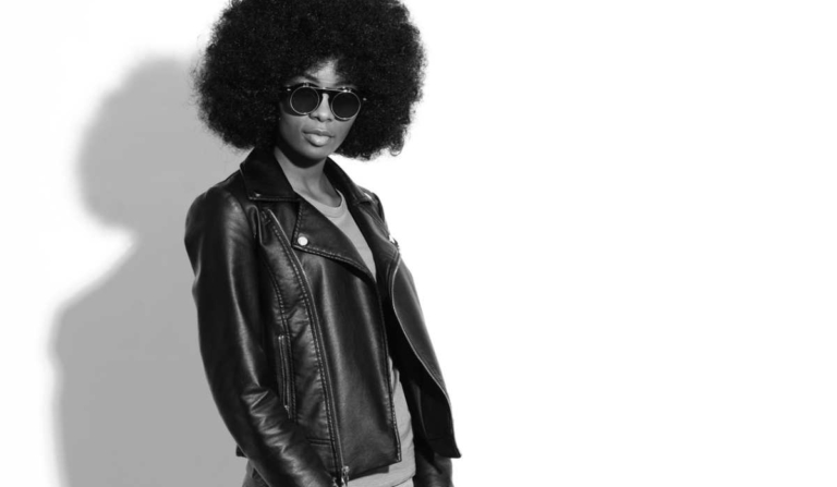 Black and white portrait of a Black woman with an afro wearing dark sunglasses, a leather jacket, a T-shirt and jeans, standing confidently and looking away from the camera, reflecting why January is such an awkward month for fashion.