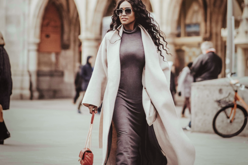 A stylish Black woman with long curls wearing a neutral midi dress and flowing light coat, walking outdoors with a red handbag in hand.