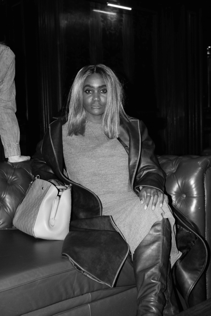 Woman seated on a leather sofa wearing a long leather coat and knee-high boots in a dramatic black-and-white setting.