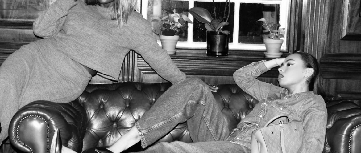 Two stylish women lounging on a leather sofa during a winter style slump, wearing layered knitwear, denim and sunglasses in a black-and-white London setting.