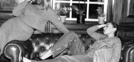 Two stylish women lounging on a leather sofa during a winter style slump, wearing layered knitwear, denim and sunglasses in a black-and-white London setting.