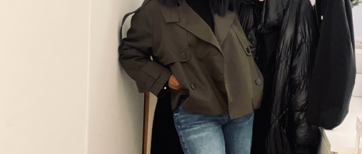 Woman wearing a deep green cropped trench coat over a black turtleneck and light blue straight-leg jeans, standing indoors beside a coat rack.