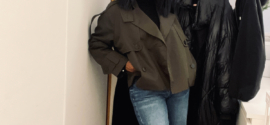 Woman wearing a deep green cropped trench coat over a black turtleneck and light blue straight-leg jeans, standing indoors beside a coat rack.