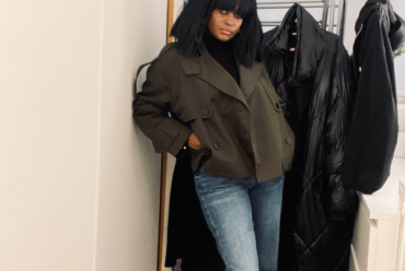 Woman wearing a deep green cropped trench coat over a black turtleneck and light blue straight-leg jeans, standing indoors beside a coat rack.