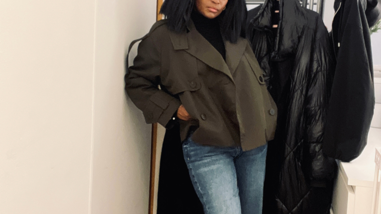 Woman wearing a deep green cropped trench coat over a black turtleneck and light blue straight-leg jeans, standing indoors beside a coat rack.