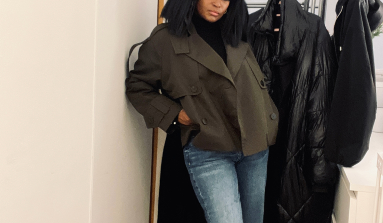 Woman wearing a deep green cropped trench coat over a black turtleneck and light blue straight-leg jeans, standing indoors beside a coat rack.