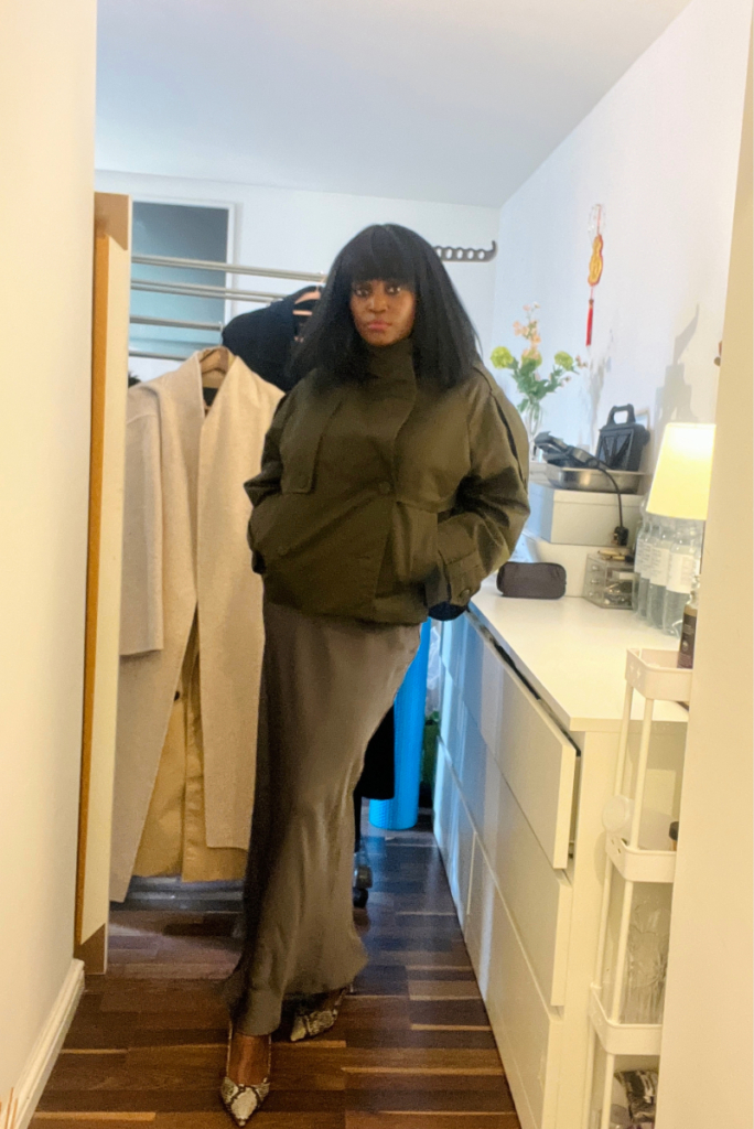 Woman wearing a deep green cropped trench coat over a dark satin midi skirt and black top, standing indoors in a narrow hallway.