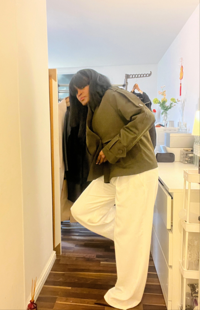 Woman wearing a dark green cropped trench coat over a black high-neck top and white wide-leg trousers, standing indoors with one leg slightly bent.