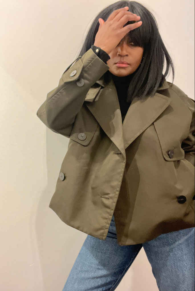 Close-up of a woman wearing a deep green cropped trench coat over a black top with blue jeans, adjusting her hair.