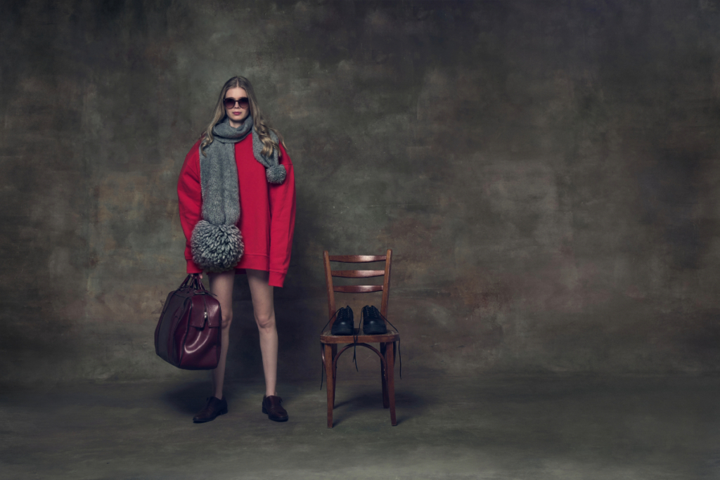 Woman wearing a red oversized sweater with a grey scarf and burgundy handbag standing beside a wooden chair.