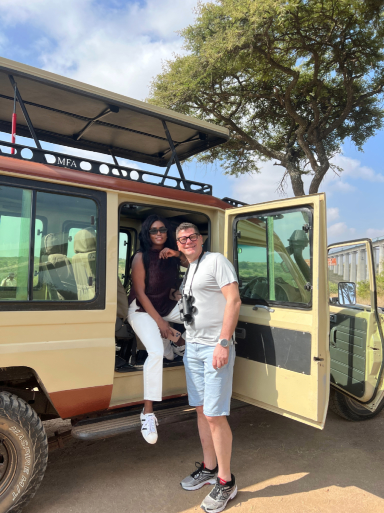 Couple standing at the open door of a safari vehicle under an acacia tree in Kenya.
