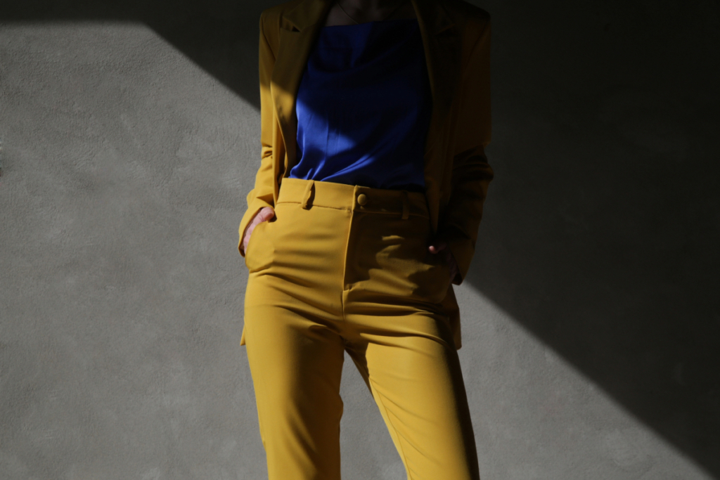 Woman wearing a mustard yellow suit with a blue blouse standing in dramatic light against a neutral wall.