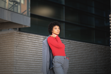 Woman wearing a red turtleneck sweater with a grey tailored suit standing outdoor.