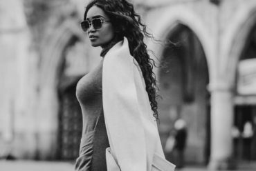 Black-and-white image of a woman in sunglasses, a grey dress and a white coat standing in a European city square