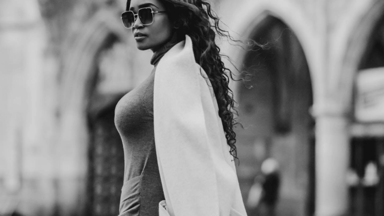 Black-and-white image of a woman in sunglasses, a grey dress and a white coat standing in a European city square