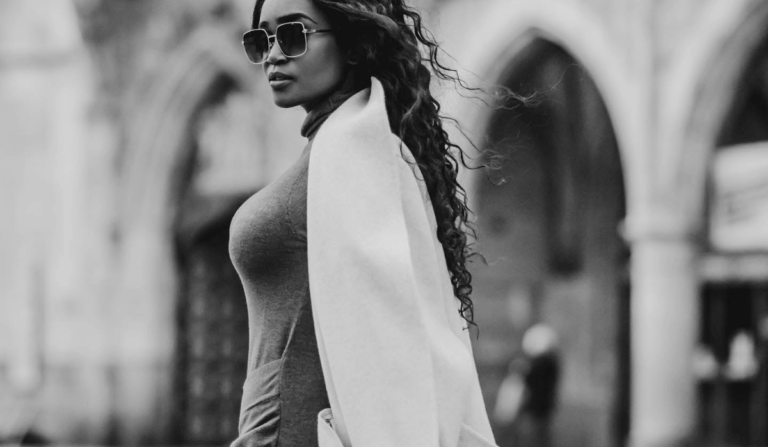 Black-and-white image of a woman in sunglasses, a grey dress and a white coat standing in a European city square