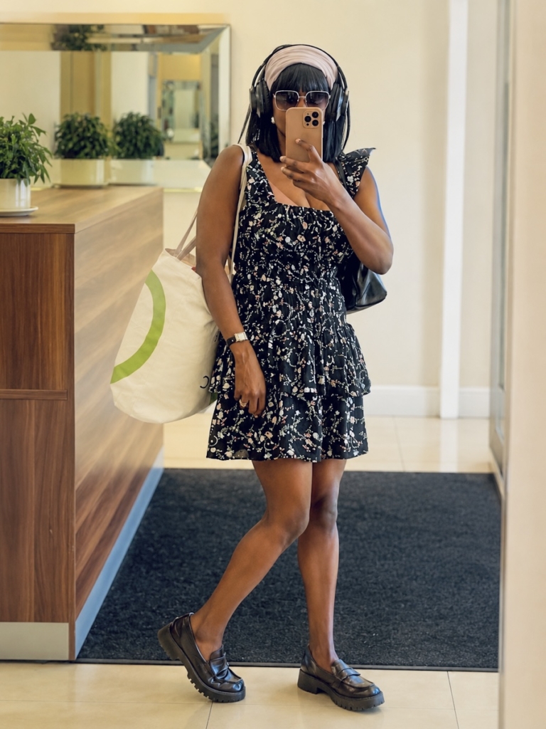 Woman wearing a black floral smocked mini dress, loafers, sunglasses and headphones while taking a mirror selfie indoors.
