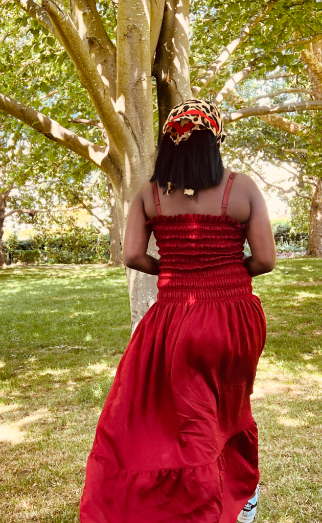 Back view of a woman wearing a red smocked maxi dress and patterned headscarf walking through a park.