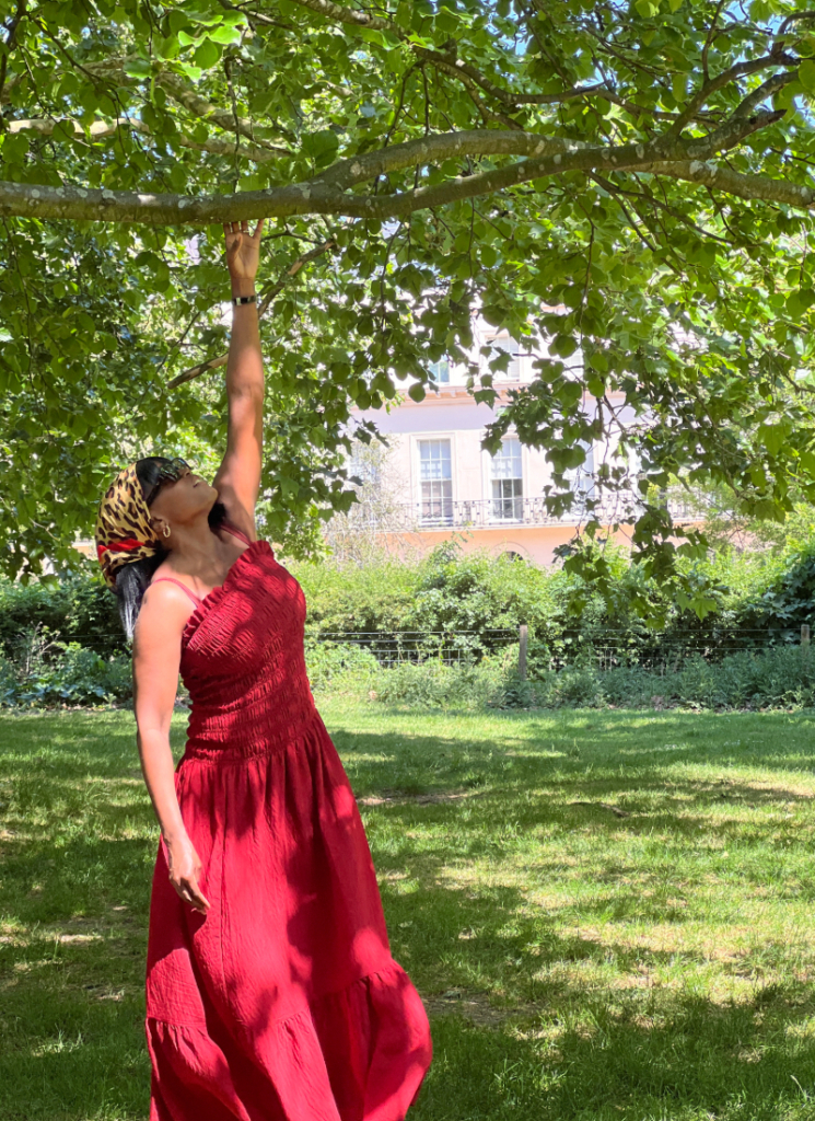 Woman wearing a red smocked maxi dress and patterned headscarf reaching towards a tree branch in a London park.