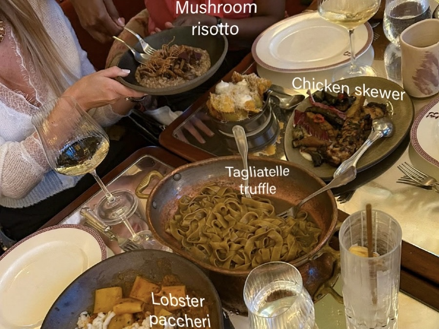 Overhead view of a restaurant table with four main courses: mushroom risotto in a dark bowl, grilled chicken skewers with vegetables, truffle tagliatelle in a copper pan, and a partially visible plate of lobster paccheri. Wine glasses and diners' hands surround the dishes.