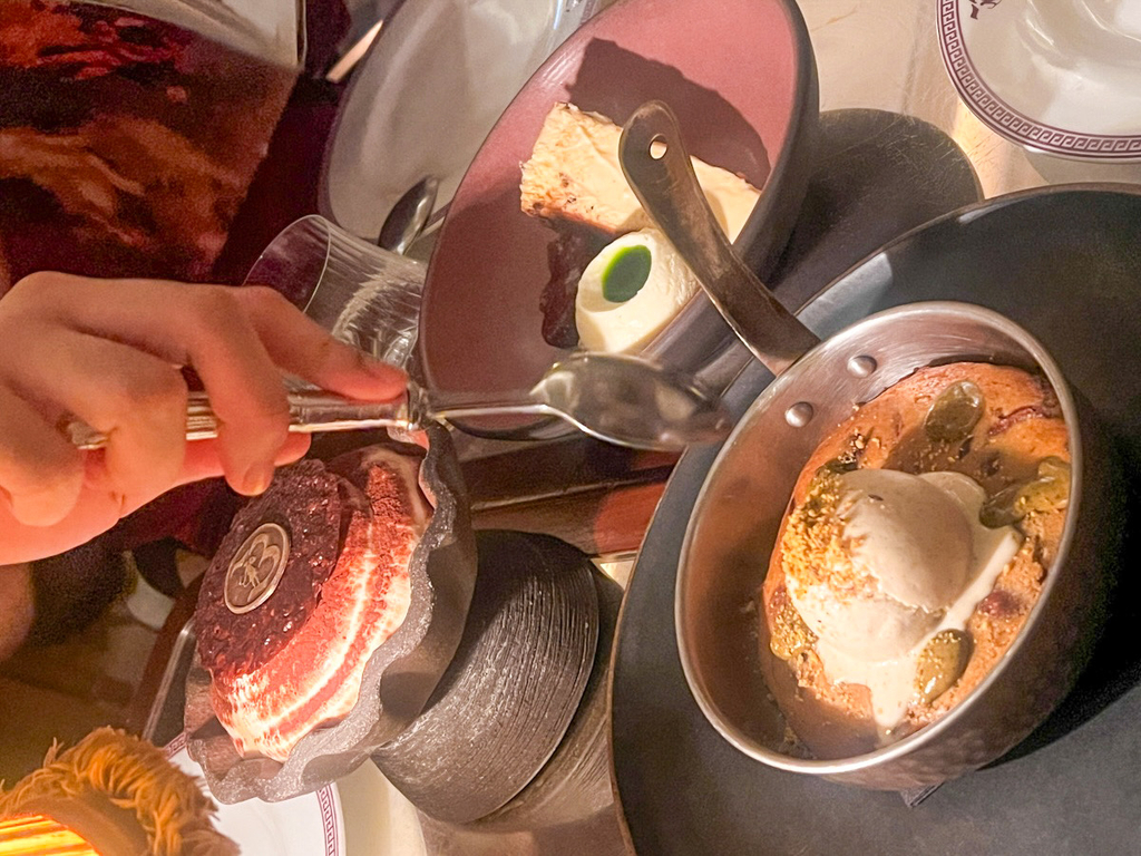 Overhead view of three desserts at Bacchanalia Mayfair: a layered dessert with a dark cocoa-dusted top on the left, a slice of cake with cream on a burgundy plate above, and a golden baked dessert topped with melting ice cream in a metal pan on the right.