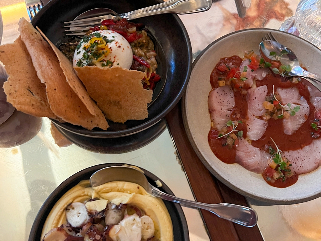 Three starters photographed from above at Bacchanalia Mayfair: a ball of burrata with vegetables and large pieces of crisp flatbread in a black bowl, thin slices of yellowtail in a red tomato dressing on a beige plate, and grilled octopus resting on a smooth yellow purée in a black bowl at the bottom of the image.